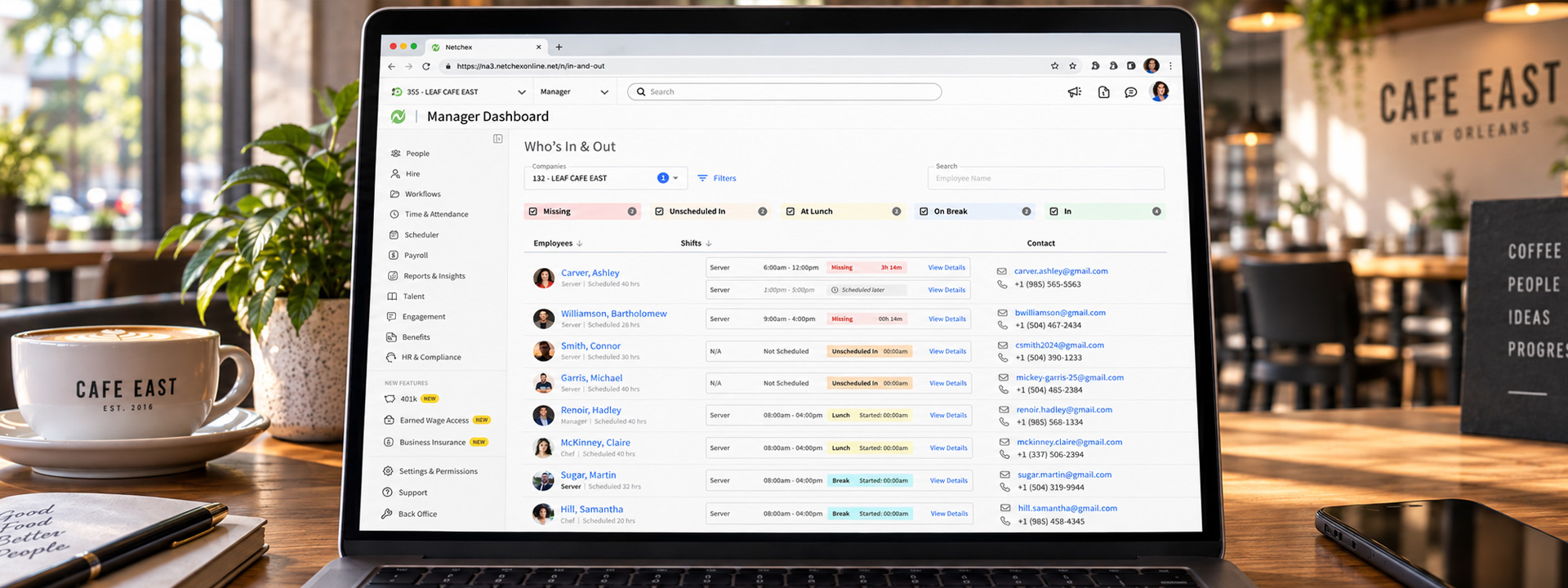 A laptop on a cafe table running the Netchex Who's In & Out manager dashboard, showing a live roster of employees with their shift statuses and contact details